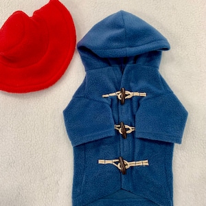 Pu&ograve; includere: Un cappotto con cappuccio in pile blu dotato di tre chiusure a levetta in legno marrone con passanti in corda beige, accompagnato da un cappello in pile rosso. Il cappotto ha maniche lunghe e cappuccio, il cappello &egrave; circolare con tesa.