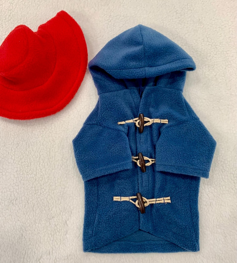 Pu&ograve; includere: Un cappotto con cappuccio in pile blu dotato di tre chiusure a levetta in legno marrone con passanti in corda beige, accompagnato da un cappello in pile rosso. Il cappotto ha maniche lunghe e cappuccio, il cappello &egrave; circolare con tesa.