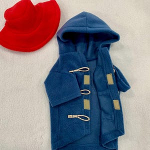Pu&ograve; includere: Un cappotto con cappuccio in pile blu dotato di tre chiusure a levetta con passanti in corda chiara e toppe rettangolari, accompagnato da un cappello da pescatore in pile rosso acceso. Entrambi gli articoli sono realizzati in pile testurizzato.