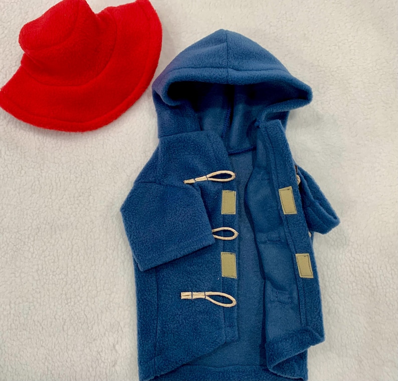 Pu&ograve; includere: Un cappotto con cappuccio in pile blu dotato di tre chiusure a levetta con passanti in corda chiara e toppe rettangolari, accompagnato da un cappello da pescatore in pile rosso acceso. Entrambi gli articoli sono realizzati in pile testurizzato.