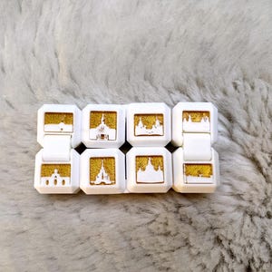 May include: A white fidget cube with eight sides, each featuring a different gold-colored image of a building. The cube is made of plastic and has a smooth, matte finish.