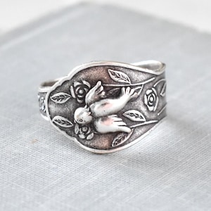 May include: A silver-toned ring with a detailed bird and floral design. The ring features a central bird motif surrounded by leaves and roses. The ring is set on a light gray surface.
