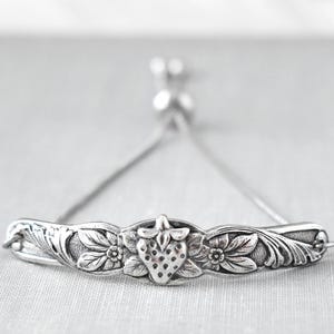 May include: Silver bracelet featuring a central strawberry motif with floral detailing. The adjustable bracelet is made of a fine chain and is set against a neutral backdrop. A charming piece of jewellery.