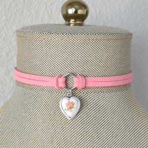 May include: A pink suede choker necklace with a silver heart charm featuring a pink rose design.