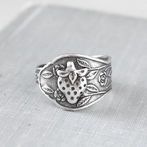 May include: A silver ring with a detailed strawberry design. The ring has a vintage aesthetic, with the strawberry at the centre, surrounded by leaves and floral accents. The ring is likely made of a metal alloy.