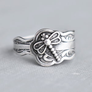 May include: A silver ring featuring a dragonfly design. The dragonfly is detailed with wings, a body, and bead-like accents.