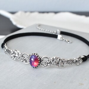 Puede incluir: Un collar gargantilla negro con un dise&ntilde;o floral plateado y una gran piedra preciosa roja y p&uacute;rpura iridiscente en el centro.