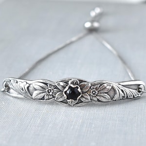 May include: Silver bracelet with a floral design and a black gemstone centre. The adjustable bracelet features a delicate chain and a decorative bar with detailed flower and leaf carvings. A stylish piece of jewellery.