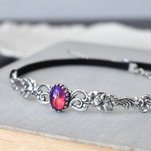 Puede incluir: Un collar gargantilla negro con un dise&ntilde;o floral plateado y una gran piedra preciosa ovalada iridiscente en el centro.