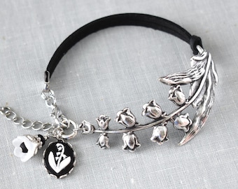 May include: Silver-toned bracelet featuring a lily of the valley design. The bracelet has a black band and a chain with a white flower charm and a black and white oval charm. The bracelet is on a light gray surface.