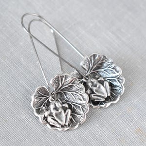 May include: Silver-toned earrings featuring a frog design. Each earring showcases a detailed frog figure resting on a textured leaf. The earrings have long, slender hooks for pierced ears. The background is a light gray surface.