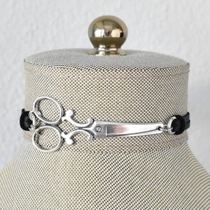 May include: A silver scissor charm choker necklace with a black band. The scissor charm is detailed and attached to the black band. The necklace is displayed on a mannequin.