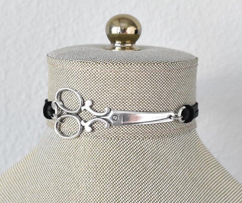 May include: A silver scissor charm choker necklace with a black band. The scissor charm is detailed and attached to the black band. The necklace is displayed on a mannequin.