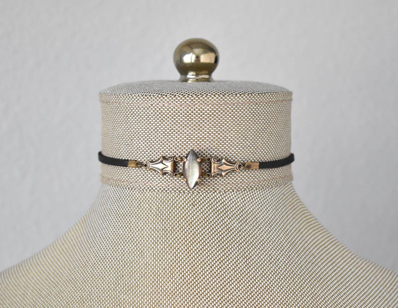 May include: A black choker necklace with a silver and gold-toned centerpiece. The centerpiece features a central oval-shaped stone and ornate metalwork. The necklace is displayed on a mannequin.