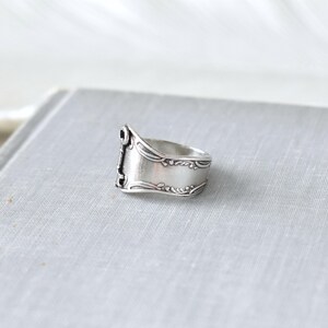 May include: A silver ring crafted from a spoon, featuring ornate detailing along the edges and a rectangular face. The ring has a vintage aesthetic, with a slightly tarnished appearance, and is set against a neutral background.
