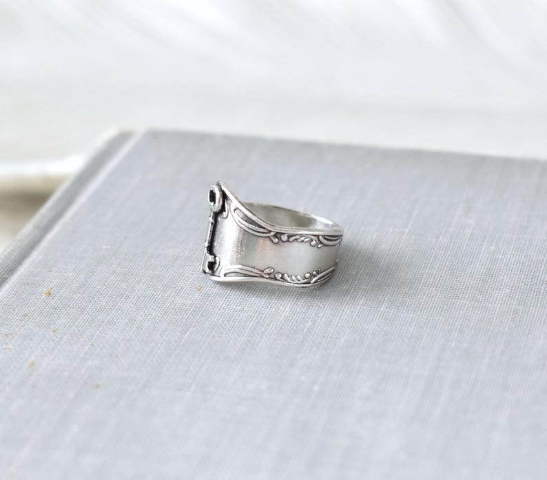 May include: A silver ring crafted from a spoon, featuring ornate detailing along the edges and a rectangular face. The ring has a vintage aesthetic, with a slightly tarnished appearance, and is set against a neutral background.