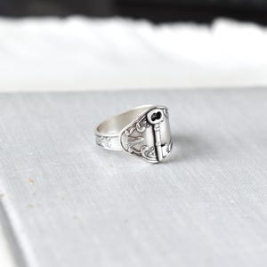 May include: A silver spoon ring with a key design. The ring features an ornate key design on the face, with decorative patterns along the band. The ring is made of a shiny, silver-colored metal.