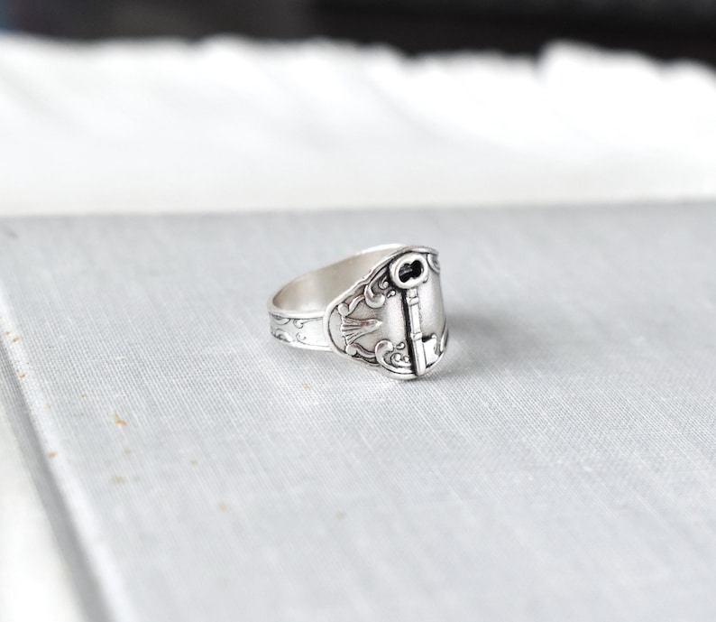 May include: A silver spoon ring with a key design. The ring features an ornate key design on the face, with decorative patterns along the band. The ring is made of a shiny, silver-colored metal.