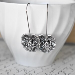 May include: Silver-toned frog earrings. Each earring features a detailed frog charm resting on a textured leaf. The earrings hang from thin, silver-colored wires, showcasing a nature-inspired design. The earrings are displayed against a white background.