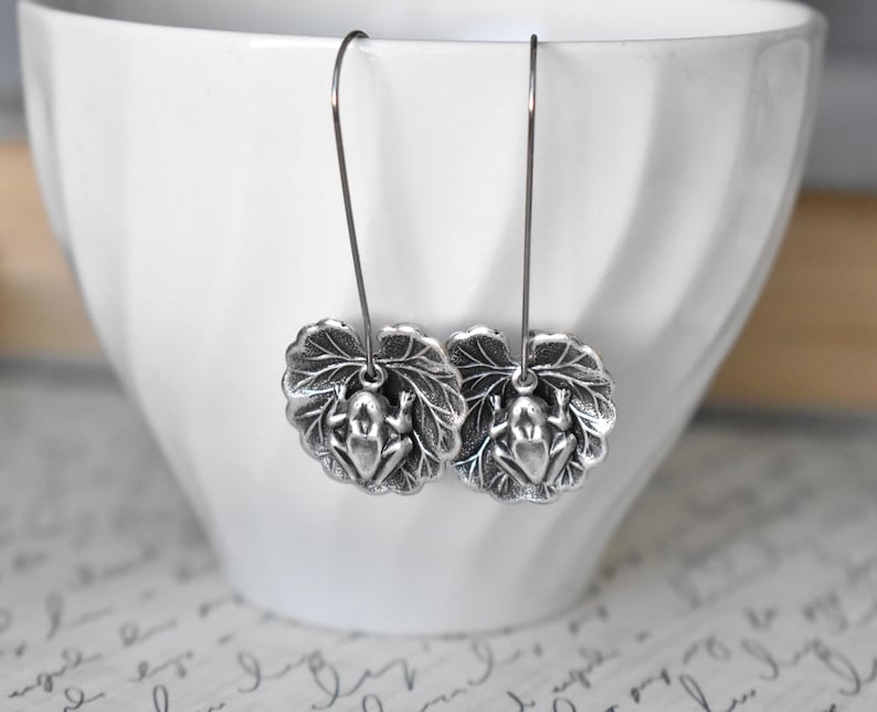 May include: Silver-toned frog earrings. Each earring features a detailed frog charm resting on a textured leaf. The earrings hang from thin, silver-colored wires, showcasing a nature-inspired design. The earrings are displayed against a white background.