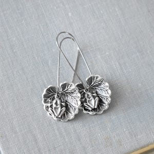 May include: A pair of silver-toned earrings featuring a frog design. Each earring has a detailed frog figure resting on a leaf-shaped base. The earrings have long, curved ear wires.