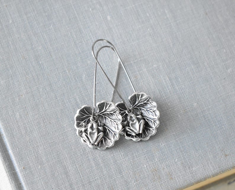 May include: A pair of silver-toned earrings featuring a frog design. Each earring has a detailed frog figure resting on a leaf-shaped base. The earrings have long, curved ear wires.