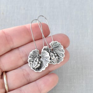May include: Silver-toned earrings featuring a frog perched on a detailed leaf design. The earrings have a long, curved hook for easy wear. The leaf and frog are intricately detailed, creating a nature-inspired accessory.