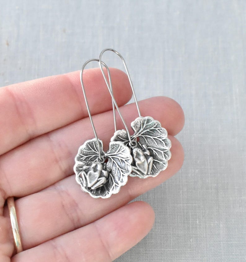 May include: Silver-toned earrings featuring a frog perched on a detailed leaf design. The earrings have a long, curved hook for easy wear. The leaf and frog are intricately detailed, creating a nature-inspired accessory.