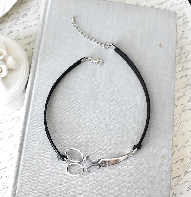 May include: A black cord necklace featuring a silver-toned scissor pendant. The scissors have detailed handles and blades. The necklace has a silver chain extender and clasp. The jewelry is displayed on a light gray book.