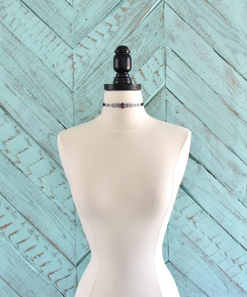 Puede incluir: Un collar gargantilla negro con una cadena plateada y un colgante de piedra roja.