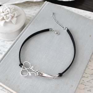 May include: A black cord bracelet featuring a silver-colored scissor charm. The scissors have detailed handles and blades. The bracelet has a lobster clasp and an extender chain for adjustable sizing. The jewelry is displayed on a light gray surface.