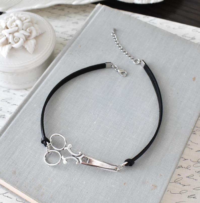 May include: A black cord bracelet featuring a silver-colored scissor charm. The scissors have detailed handles and blades. The bracelet has a lobster clasp and an extender chain for adjustable sizing. The jewelry is displayed on a light gray surface.