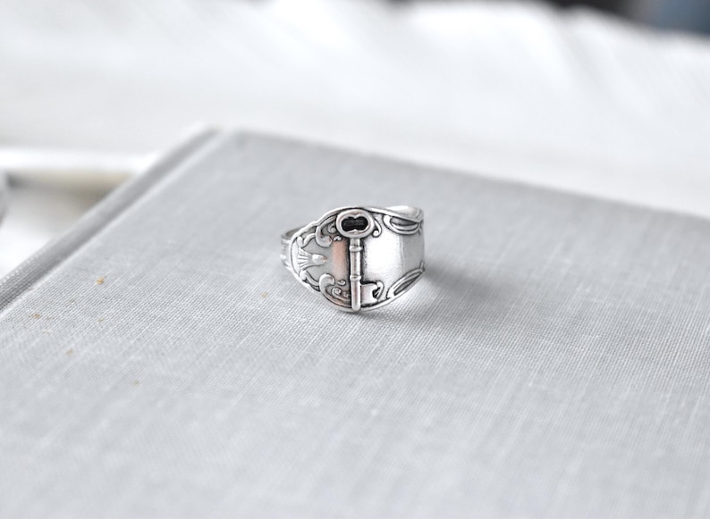 May include: A silver-toned ring with a key design. The ring features an ornate key in the center, with decorative patterns around the edges. The ring is set against a light gray background.