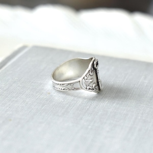 May include: A silver ring with intricate floral and bird designs. The ring has a wide band with detailed patterns and a decorative element on the face. The ring is set against a light gray background.