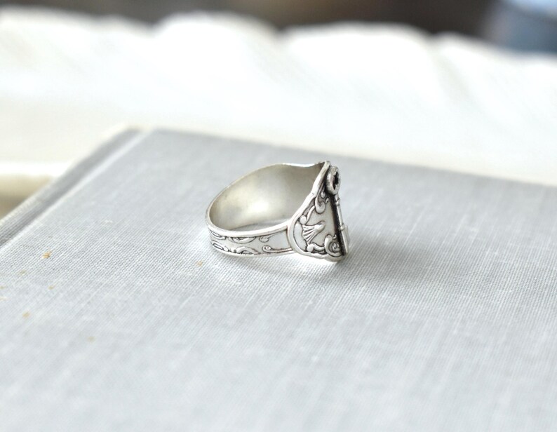 May include: A silver ring with intricate floral and bird designs. The ring has a wide band with detailed patterns and a decorative element on the face. The ring is set against a light gray background.
