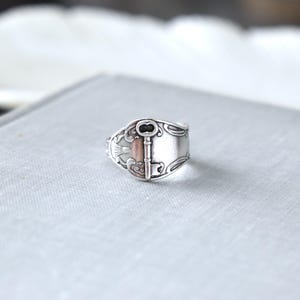 May include: A silver-toned ring with a key design. The ring features a rectangular face with a key motif and decorative flourishes. The key's details are highlighted with a darker tone, adding contrast to the silver metal.