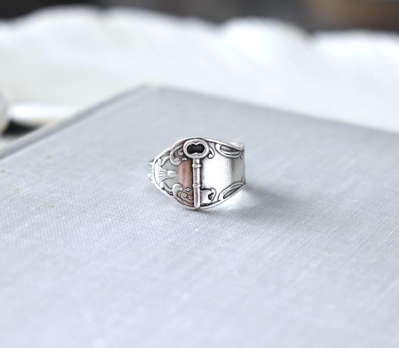 May include: A silver-toned ring with a key design. The ring features a rectangular face with a key motif and decorative flourishes. The key's details are highlighted with a darker tone, adding contrast to the silver metal.