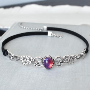 Puede incluir: Un collar choker plateado con una cinta de terciopelo negro y un dise&ntilde;o floral con una gran piedra preciosa iridiscente p&uacute;rpura y rosa.