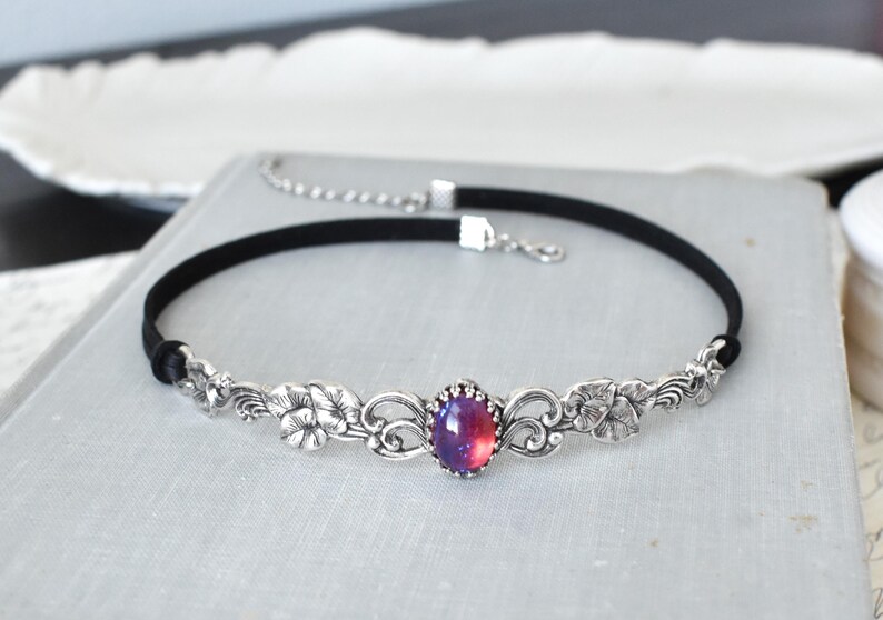 Puede incluir: Un collar choker plateado con una cinta de terciopelo negro y un dise&ntilde;o floral con una gran piedra preciosa iridiscente p&uacute;rpura y rosa.