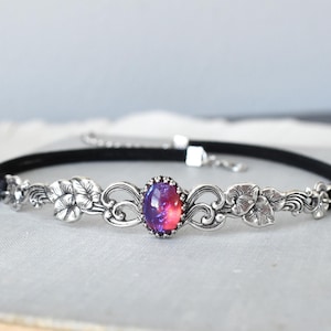 Puede incluir: Un collar gargantilla de terciopelo negro con un dise&ntilde;o floral plateado y una piedra de &oacute;palo p&uacute;rpura y rosa en el centro.