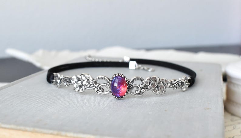 Puede incluir: Un collar gargantilla de terciopelo negro con un dise&ntilde;o floral plateado y una piedra de &oacute;palo p&uacute;rpura y rosa en el centro.