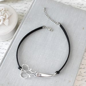 May include: A black choker necklace featuring a silver scissor pendant. The scissors are detailed with intricate designs and attached to a black cord. The necklace has a silver chain for adjustable sizing.