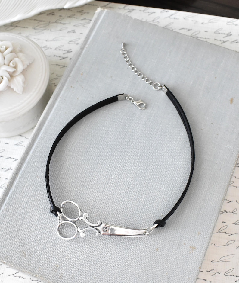 May include: A black choker necklace featuring a silver scissor pendant. The scissors are detailed with intricate designs and attached to a black cord. The necklace has a silver chain for adjustable sizing.
