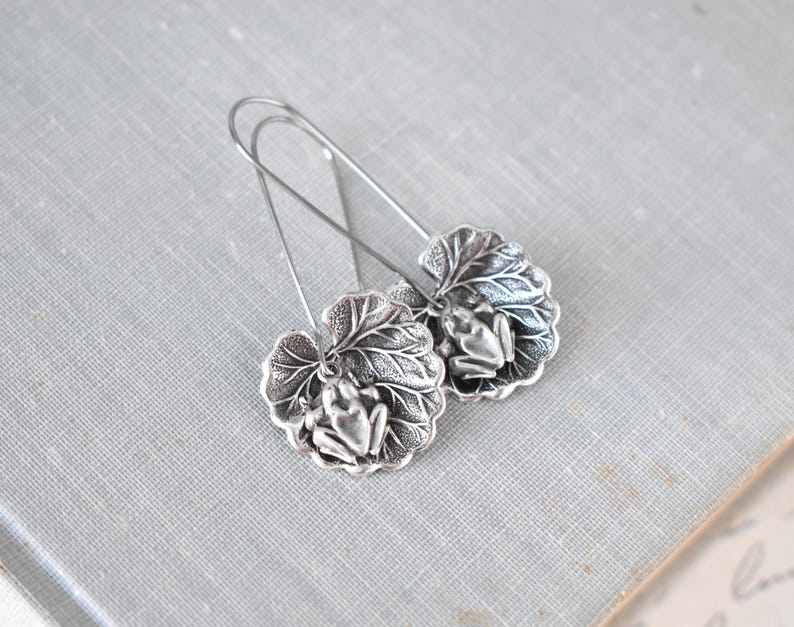 May include: Silver-toned earrings featuring a frog design. Each earring showcases a detailed frog figure resting on a textured leaf. The earrings have long, slender hooks for pierced ears. The background is a light gray surface.