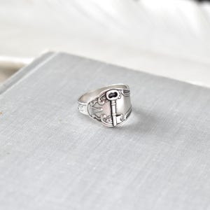 May include: A silver-colored ring with a key design. The ring has an ornate, vintage style with a key-shaped centerpiece and decorative detailing. The ring is set on a light gray surface.