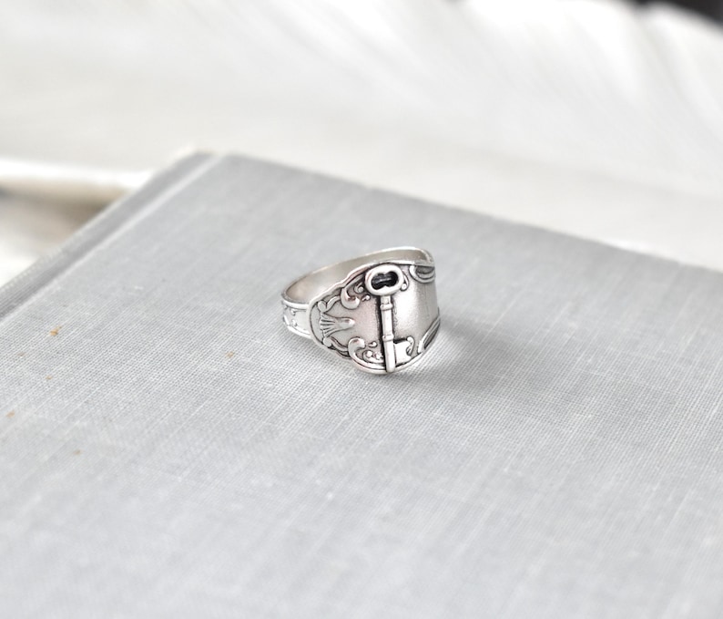 May include: A silver-colored ring with a key design. The ring has an ornate, vintage style with a key-shaped centerpiece and decorative detailing. The ring is set on a light gray surface.