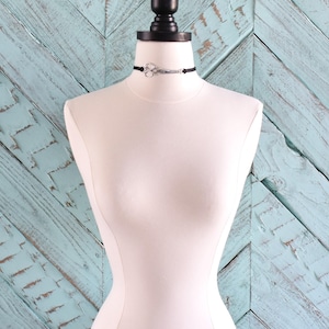 May include: A black choker necklace with a silver scissors pendant displayed on a white mannequin. The mannequin is set against a light blue wooden background. The scissors pendant is the focal point of the necklace.