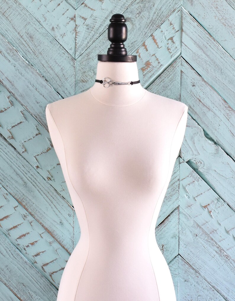 May include: A black choker necklace with a silver scissors pendant displayed on a white mannequin. The mannequin is set against a light blue wooden background. The scissors pendant is the focal point of the necklace.