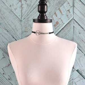 May include: A black choker necklace with a silver scissor pendant. The necklace is displayed on a white mannequin against a light blue wooden background. The scissor pendant is detailed and adds a unique touch to the accessory.