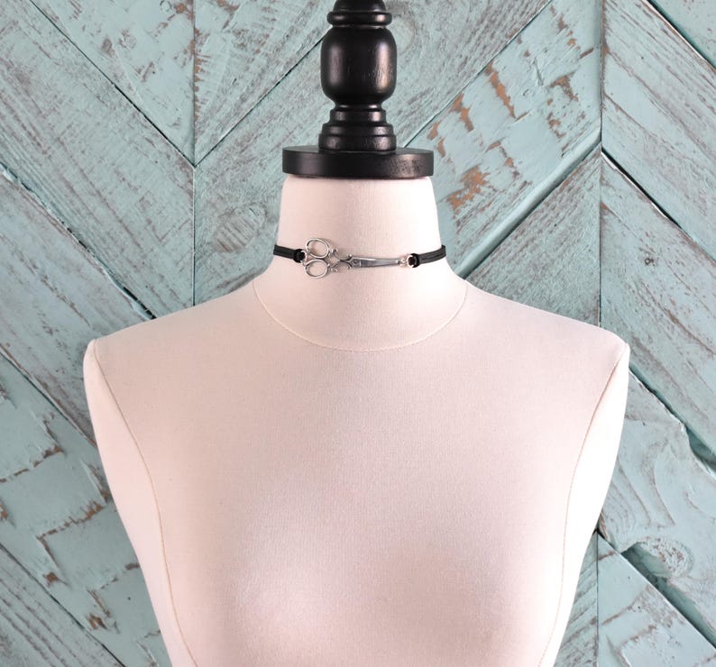 May include: A black choker necklace with a silver scissor pendant. The necklace is displayed on a white mannequin against a light blue wooden background. The scissor pendant is detailed and adds a unique touch to the accessory.